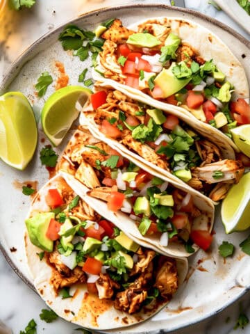 Pulled chicken tacos in tortillas with chopped tomatoes and avocado on a white plate.