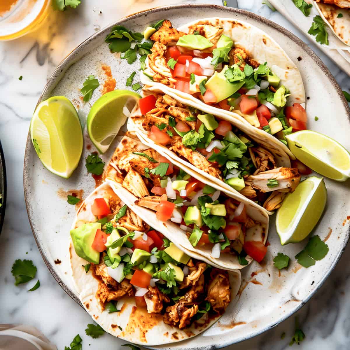 Pulled chicken tacos in tortillas with chopped tomatoes and avocado on a white plate.